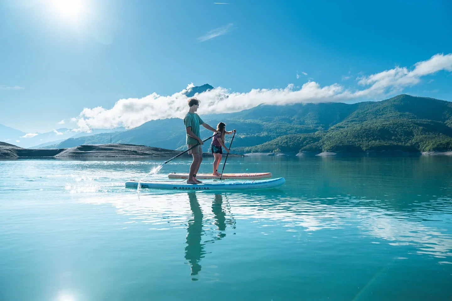Dağ manzaralı gölde iki kişi Aqua Marina stand up paddle sörfü yapıyor