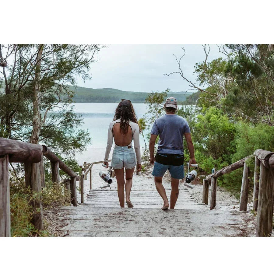 Göl manzaralı ormanda, elinde sualtı scooterı tutan genç çift ahşap merdivenden sahile iniyor