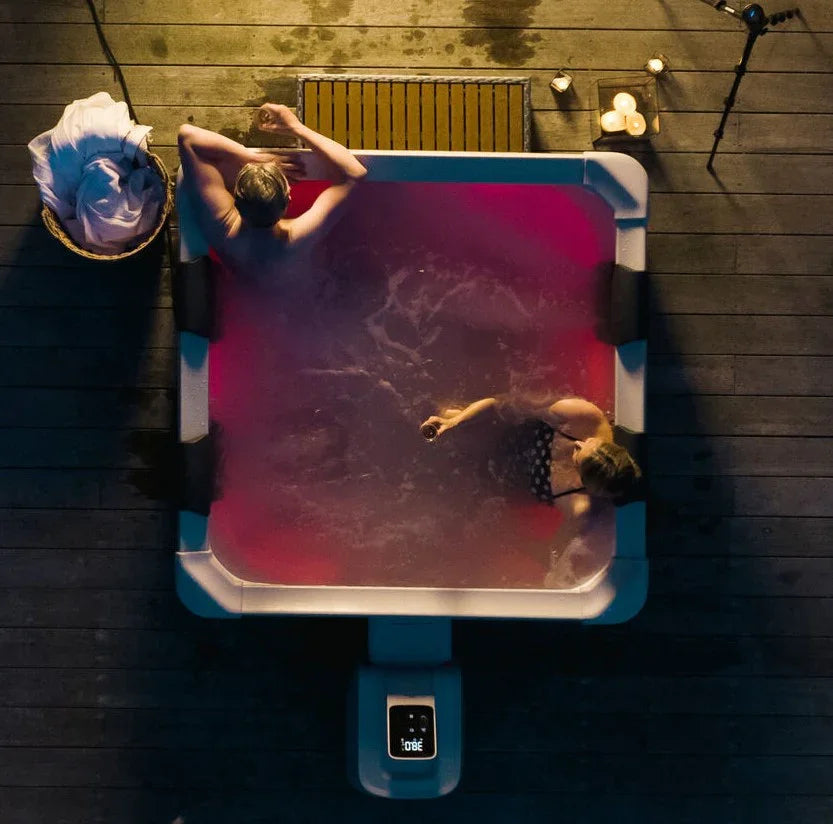 Ahşap zemin üzerinde iki kişinin içinde bulunduğu kırmızı ışıklı kare spa küveti ve çevresinde mumlar