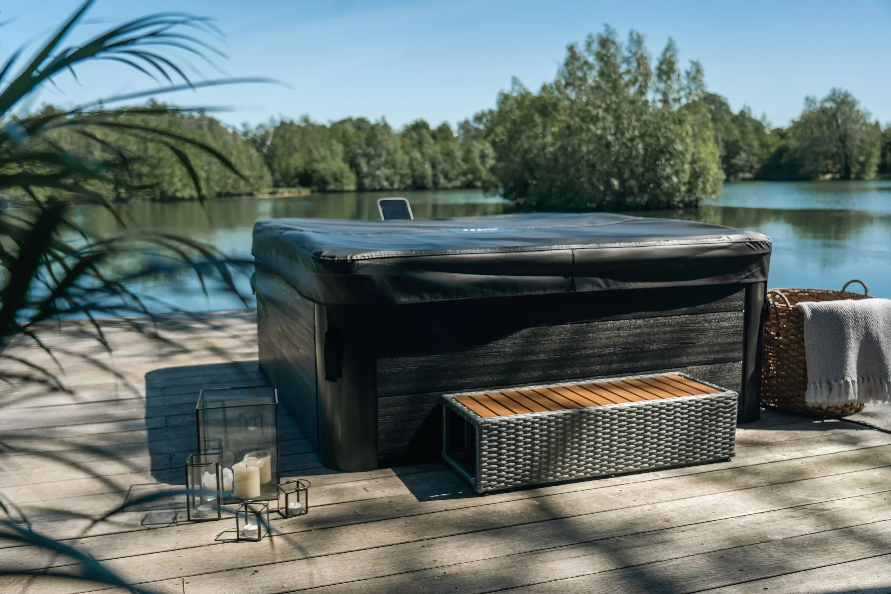 Dış mekanda göl kenarında kapaklı siyah jakuzili teras, rattan oturma bankı ve dekoratif mumluklar