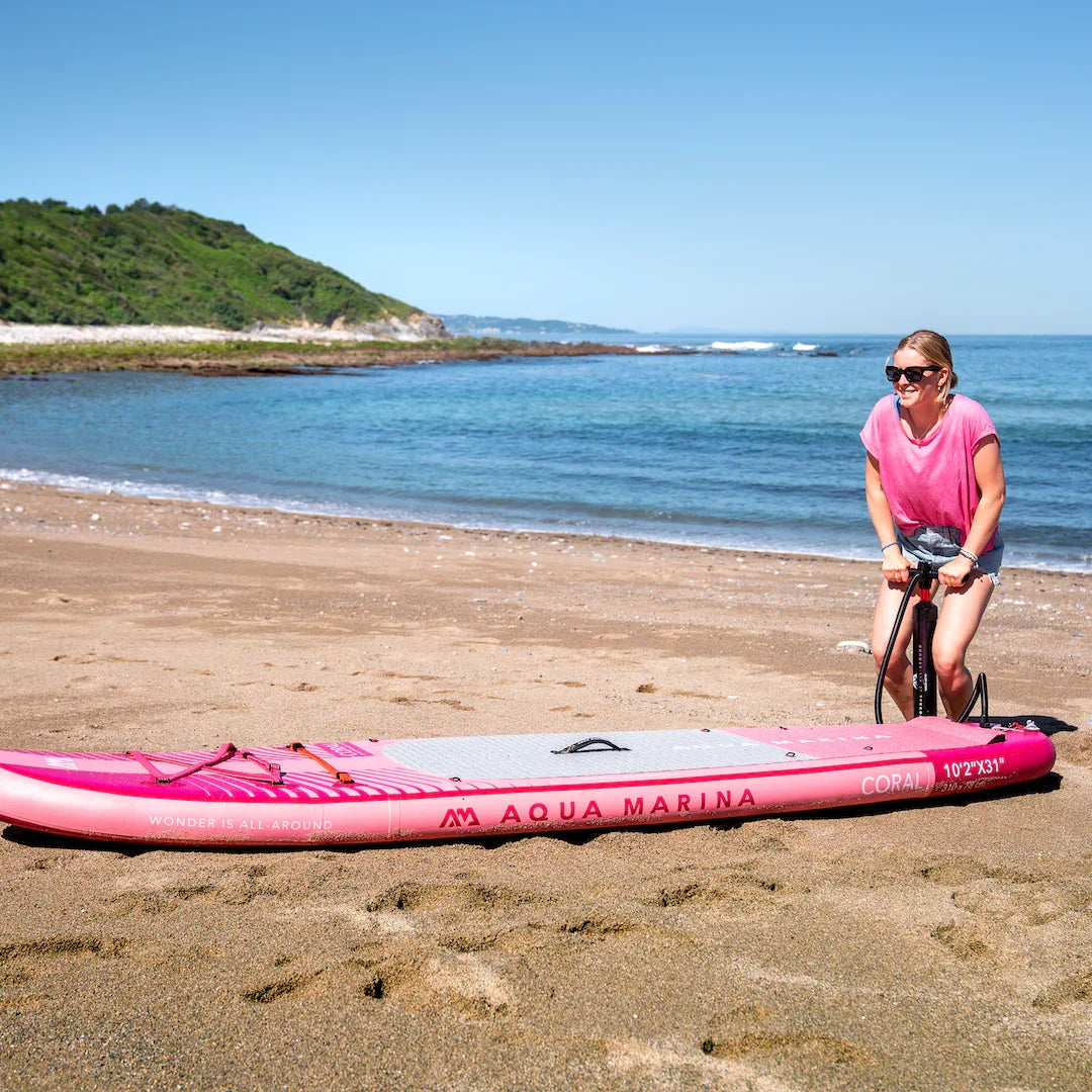Woman inflating pink Aqua Marina Coral inflatable stand up paddleboard on sandy beach by ocean