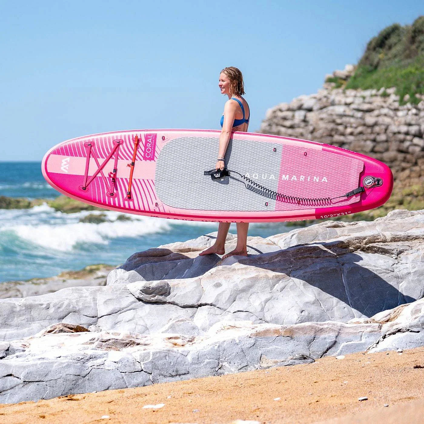 Woman carrying pink Aqua Marina Coral stand-up paddleboard on rocky beach by ocean
