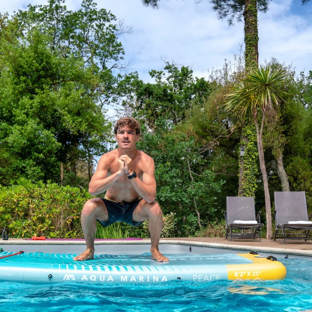 Suda duran Aqua Marina Peace sörf tahtasında çömelme pozisyonunda erkek sporcu, havuz ve yeşil ağaçlar arka planda