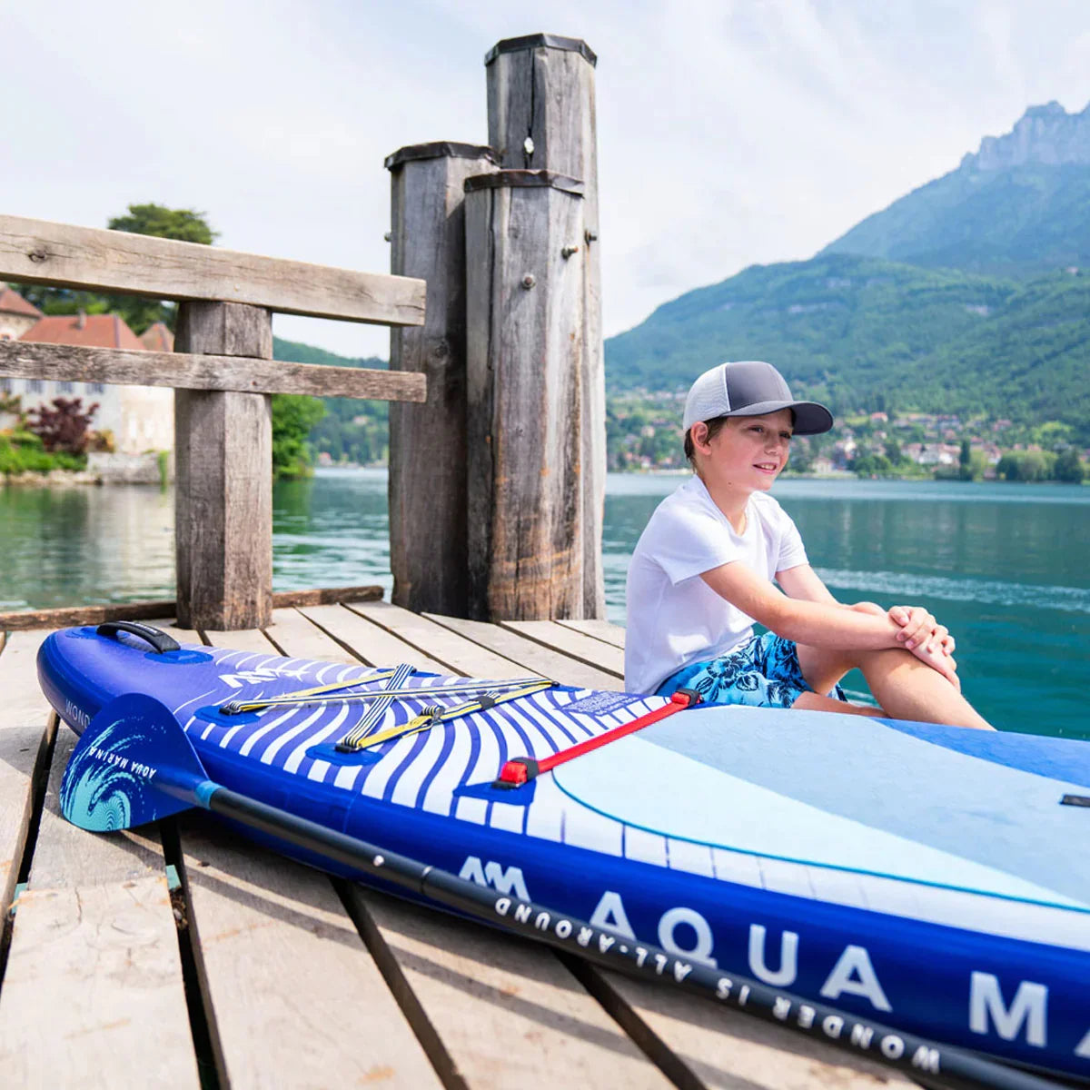 Mavi Aqua Marina stand up paddle board ve iskelede oturan şapkalı çocuk, dağ manzaralı göl kenarı